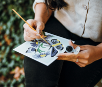 Person painting a butterfly design on a piece of paper with a paintbrush.