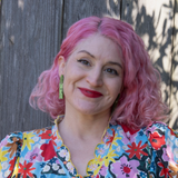 Person with pink hair wearing a colorful floral shirt against a wooden fence.
