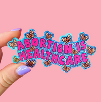 Sticker with 'ABORTION IS HEALTHCARE' text held by a hand against a pink background