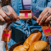Person holding a small wooden box with a pink label, surrounded by bread in a basket.
