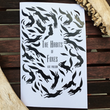 Book cover of 'The Habits of Foxes' with black fox illustrations on a white background, displayed on a wooden surface.