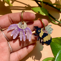 Hand holding two earrings, one purple flower and one bee design, with green leaves in the background.