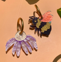 Two earrings, one with a purple flower and gold base, the other with a bee design, on a beige background with green leaves.