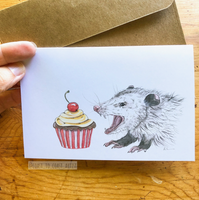 Card with an illustration of an opossum and a cupcake, held over a wooden surface with a brown envelope.