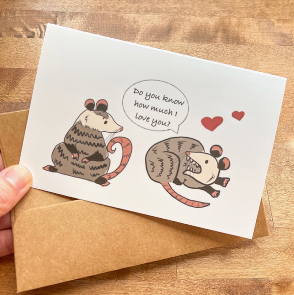 Greeting card with two illustrated opossums and a speech bubble on a wooden surface
