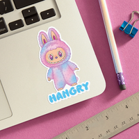 Sticker with a character named 'HANGRY' on a pink surface with a laptop, pencil, and ruler.