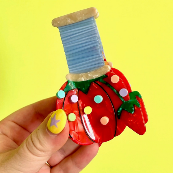 Hand holding a decorative spool of thread shaped like a strawberry against a yellow background
