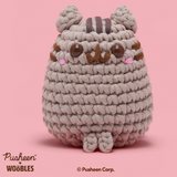 Crocheted cat toy with striped hat on a pink background, featuring the Pusheen brand.