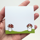 Hand holding a small white notepad with cartoon mushrooms on a grassy background